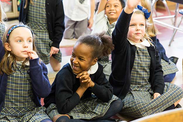 A diverse group of children sitting in a classroom, eagerly raising their hands to participate in a discussion.