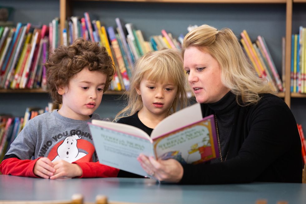 Female teacher reading book to 2 onlooking students.
